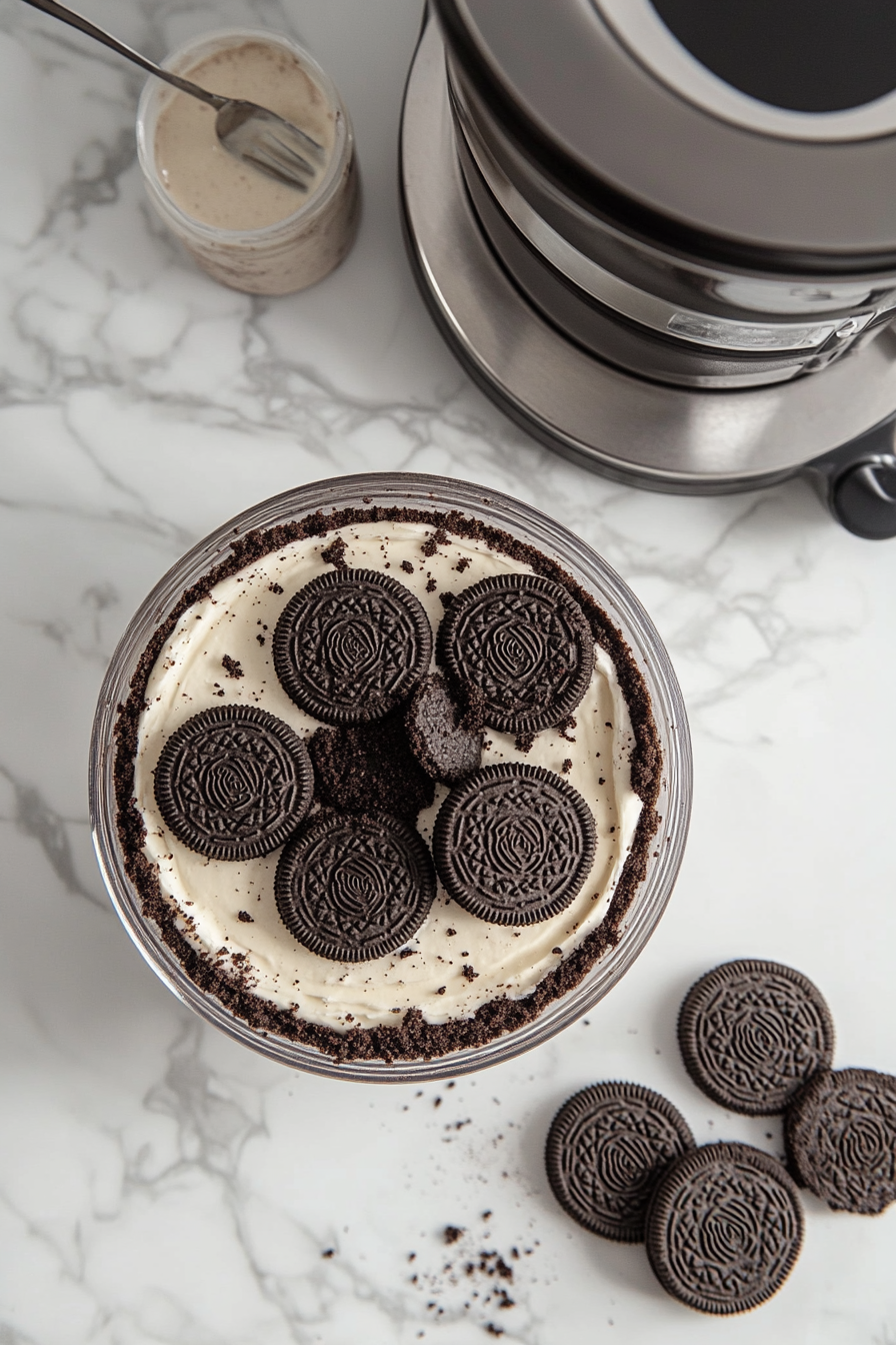 A 9-inch springform pan sits on the white marble cooktop, lightly greased and lined with parchment paper. A package of Oreos is placed beside the pan, with the cookies being crushed into fine crumbs using a food processor. The crumbs are mixed with melted butter and are being pressed evenly into the bottom and sides of the pan, forming a solid crust base.