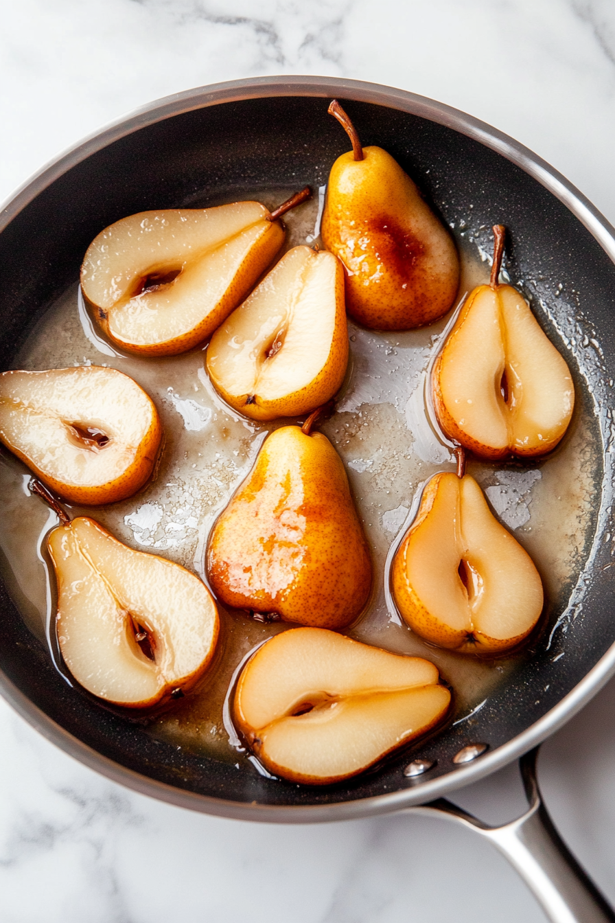 Halved pears placed cut side down in a black skillet on the white marble cooktop. The pears are beginning to caramelize, with the edges of the pan glistening with melted butter