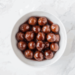 Top-down view of sweet and savory meatballs cooked in a slow cooker using frozen meatballs, ketchup, and grape jelly. The meatballs are served in a white bowl, glistening with thick, tangy red-brown sauce. The image highlights the finished dish on a white marble countertop, showcasing the richness and appeal of this classic party appetizer.