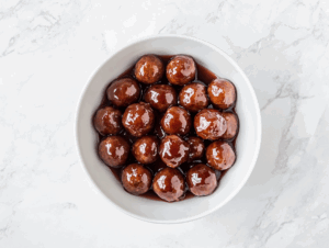 Top-down view of sweet and savory meatballs cooked in a slow cooker using frozen meatballs, ketchup, and grape jelly. The meatballs are served in a white bowl, glistening with thick, tangy red-brown sauce. The image highlights the finished dish on a white marble countertop, showcasing the richness and appeal of this classic party appetizer.