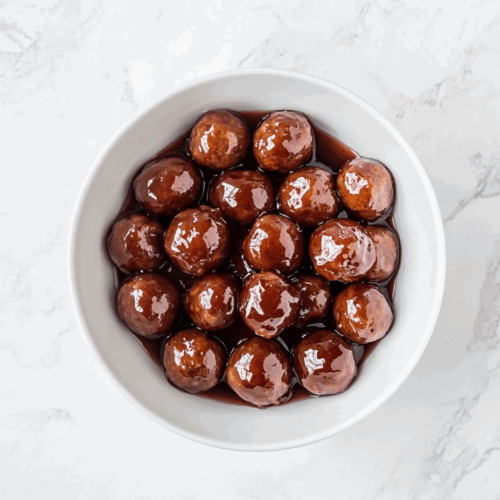 Top-down view of sweet and savory meatballs cooked in a slow cooker using frozen meatballs, ketchup, and grape jelly. The meatballs are served in a white bowl, glistening with thick, tangy red-brown sauce. The image highlights the finished dish on a white marble countertop, showcasing the richness and appeal of this classic party appetizer.
