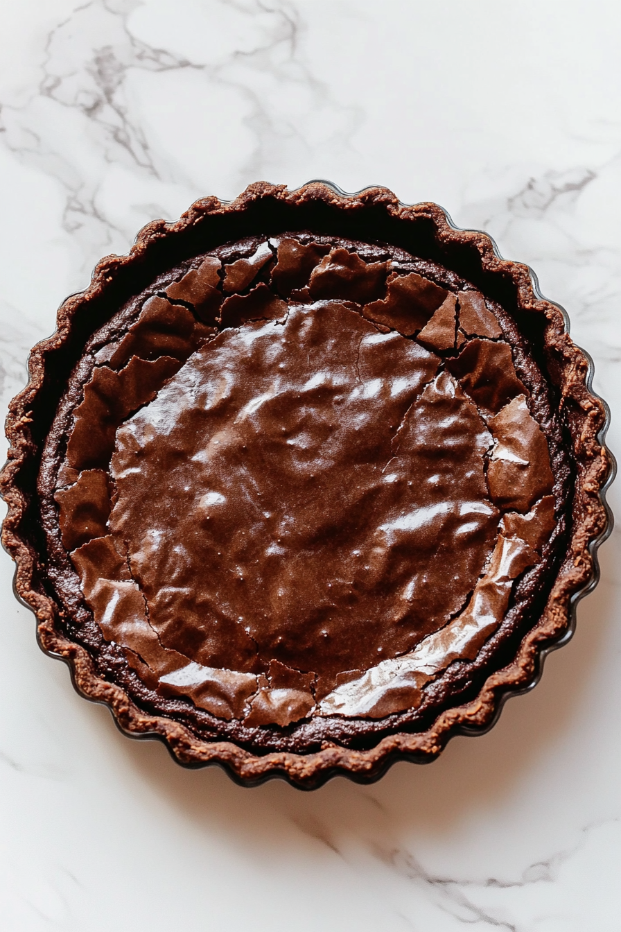 A close-up of a chocolate brownie pie in a tart tin on a white marble countertop, just after being gently tapped to settle the brownie layer. The surface has slight ripples, with a glossy, rich chocolate finish.