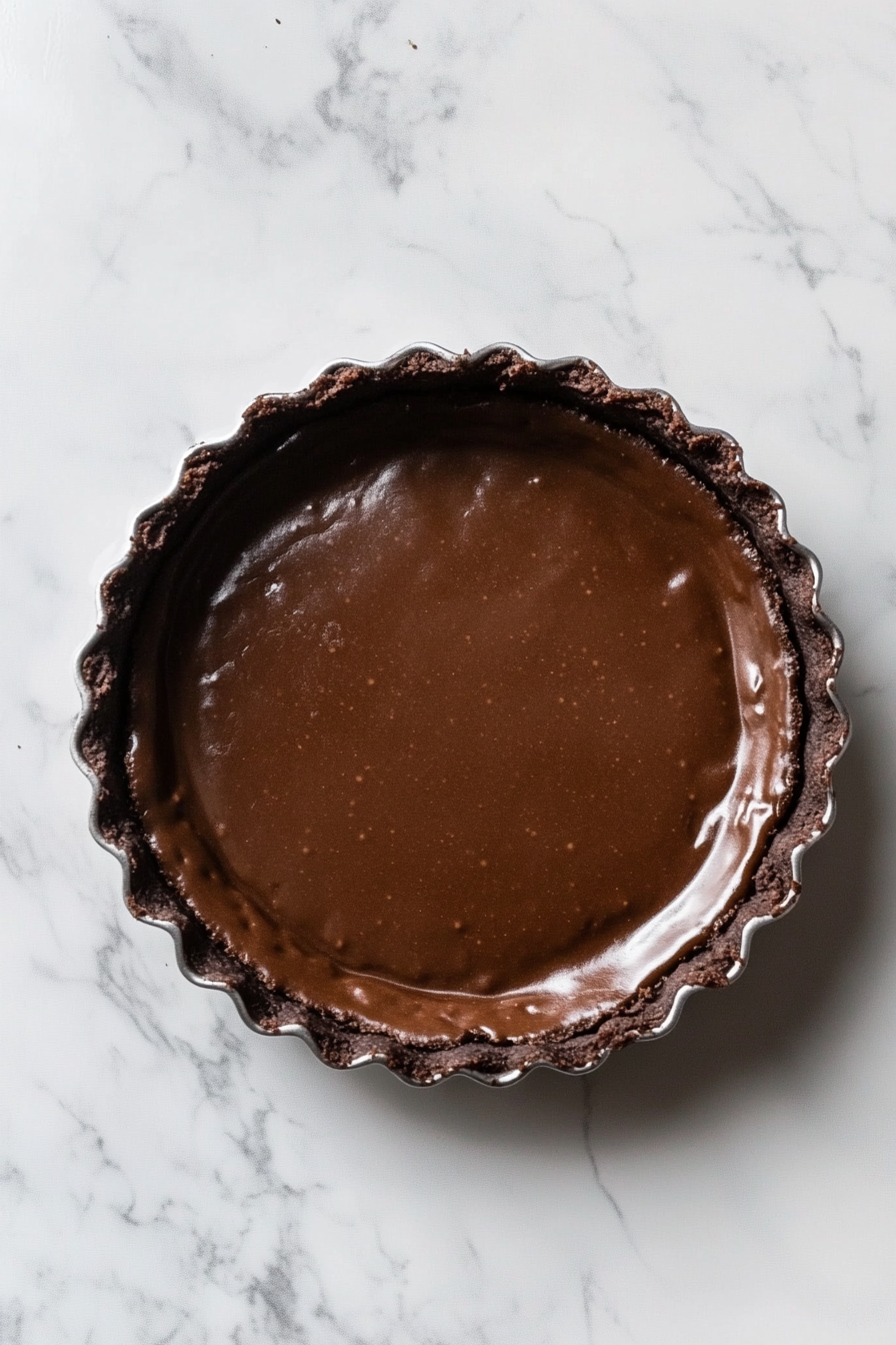 A close-up of a 9-inch tart tin on a white marble countertop showing the partially baked crust. The surface is firm and slightly darkened, with the fluted edges retaining their neat shape, ready for the next layer.