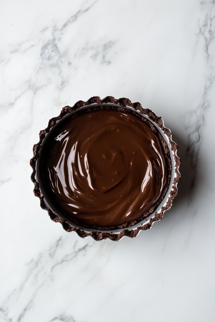 A close-up of a 9-inch tart tin on a white marble countertop, filled with a partially baked chocolate crust. The thick brownie batter is being evenly spread over the crust, creating a smooth, glossy surface.