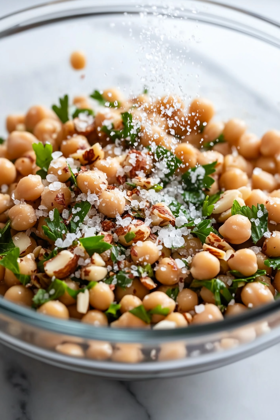 Close-up shot of a glass bowl on the white marble cooktop with the mixed almond/chickpea salad, being sprinkled with salt and freshly cracked black pepper from above.