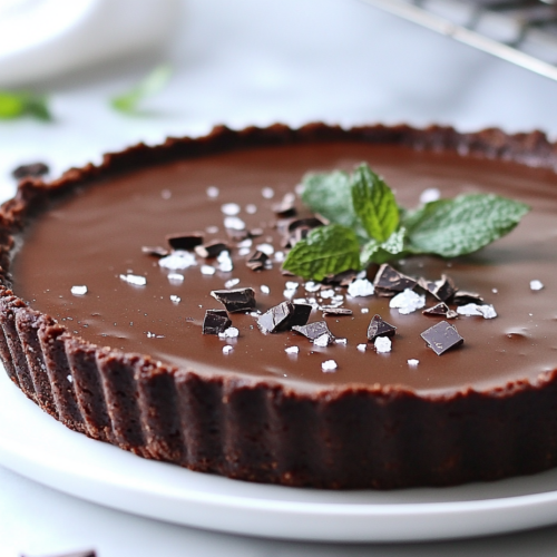 A close-up of a slice of vegan dark chocolate tart served on a white ceramic plate on a white marble countertop. The slice has a smooth, glossy chocolate filling topped with flaky sea salt and rests on a rich cocoa crust. The plate is adorned with cocoa nibs and fresh mint leaves, while the rest of the tart is visible in the background on a wire rack.