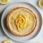 A close-up of a perfectly set vegan lemon pie on a white ceramic plate, garnished with spirals of freshly grated lemon zest artfully arranged on its smooth, creamy surface. The vibrant yellow filling contrasts with the crisp, golden cookie crust, while the plate sits on a white marble countertop surrounded by scattered lemon slices and zest.