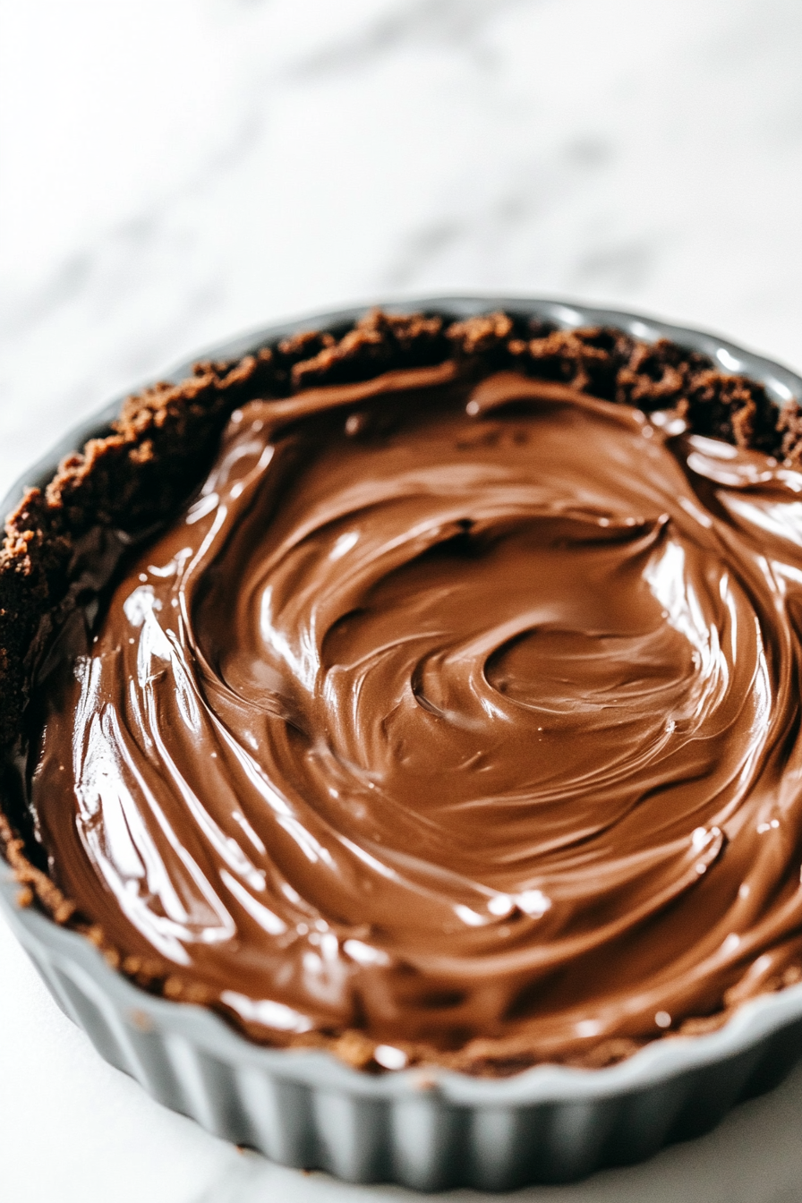 A close-up of a tart pan on a white marble countertop, featuring the glossy chocolate filling spread evenly over the crust with a perfectly smooth and shiny surface.