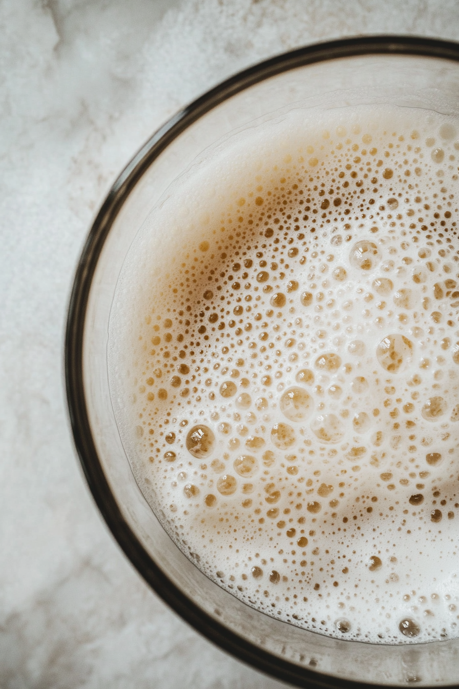 A close-up of a glass mixing bowl on a white marble countertop filled with whipped aquafaba. The mixture is thick, airy, and glossy, with small evenly dispersed bubbles, showcasing the light, cloud-like texture.