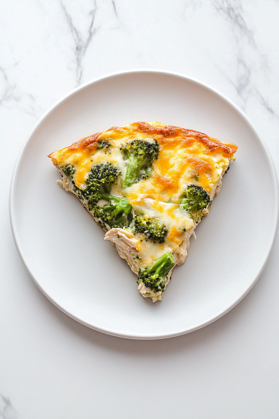 A slice of Keto Chicken Broccoli Casserole is served on a white plate, revealing layers of creamy cheese, tender chicken, and bright green broccoli. The golden, bubbly cheese topping is intact, with the plate placed on the white marble cooktop for a clean and minimal presentation