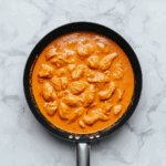 Top-down view of a rich and creamy butter chicken dish served in a large black skillet. The thick tomato and cream-based gravy surrounds tender chicken chunks, making the dish look warm, comforting, and flavorful. The white marble countertop and simple background highlight the orange-colored curry, creating an inviting presentation of this classic Indian dish.