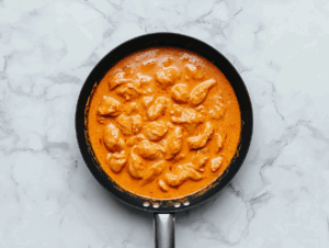 Top-down view of a rich and creamy butter chicken dish served in a large black skillet. The thick tomato and cream-based gravy surrounds tender chicken chunks, making the dish look warm, comforting, and flavorful. The white marble countertop and simple background highlight the orange-colored curry, creating an inviting presentation of this classic Indian dish.