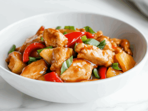 Top-down image of a colorful chicken stir-fry dish served in a shallow white bowl placed on a white marble countertop. The stir-fry features golden seared chicken breast strips mixed with red bell pepper cubes, scallion pieces, and juicy pineapple chunks, all coated in a glossy sauce. The presentation is simple and appetizing, with a vibrant, homemade feel that highlights the freshness and flavors of the ingredients used throughout the cooking process.