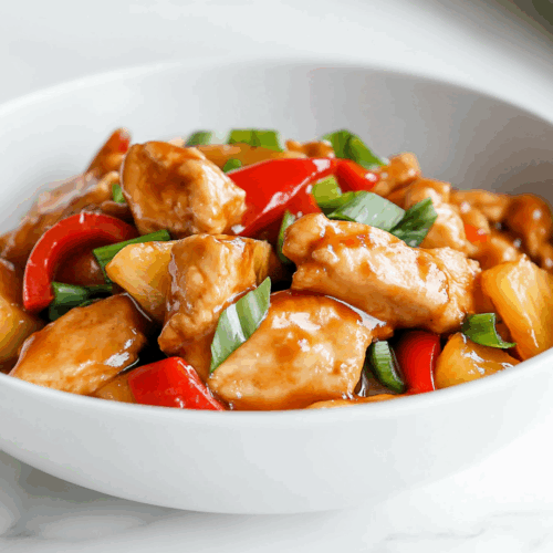 Top-down image of a colorful chicken stir-fry dish served in a shallow white bowl placed on a white marble countertop. The stir-fry features golden seared chicken breast strips mixed with red bell pepper cubes, scallion pieces, and juicy pineapple chunks, all coated in a glossy sauce. The presentation is simple and appetizing, with a vibrant, homemade feel that highlights the freshness and flavors of the ingredients used throughout the cooking process.