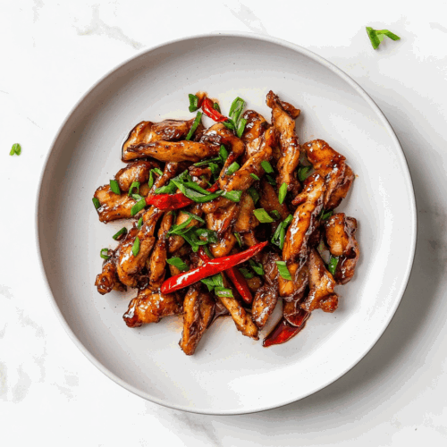 A delicious plate of crispy chilli chicken coated in a glossy, sticky sauce made from soy sauce, honey, and sriracha. The chicken is garnished with fresh chopped spring onions and served on a white ceramic plate placed on a clean white marble countertop, capturing the rich texture and mouthwatering appeal of this Indo-Chinese dish.