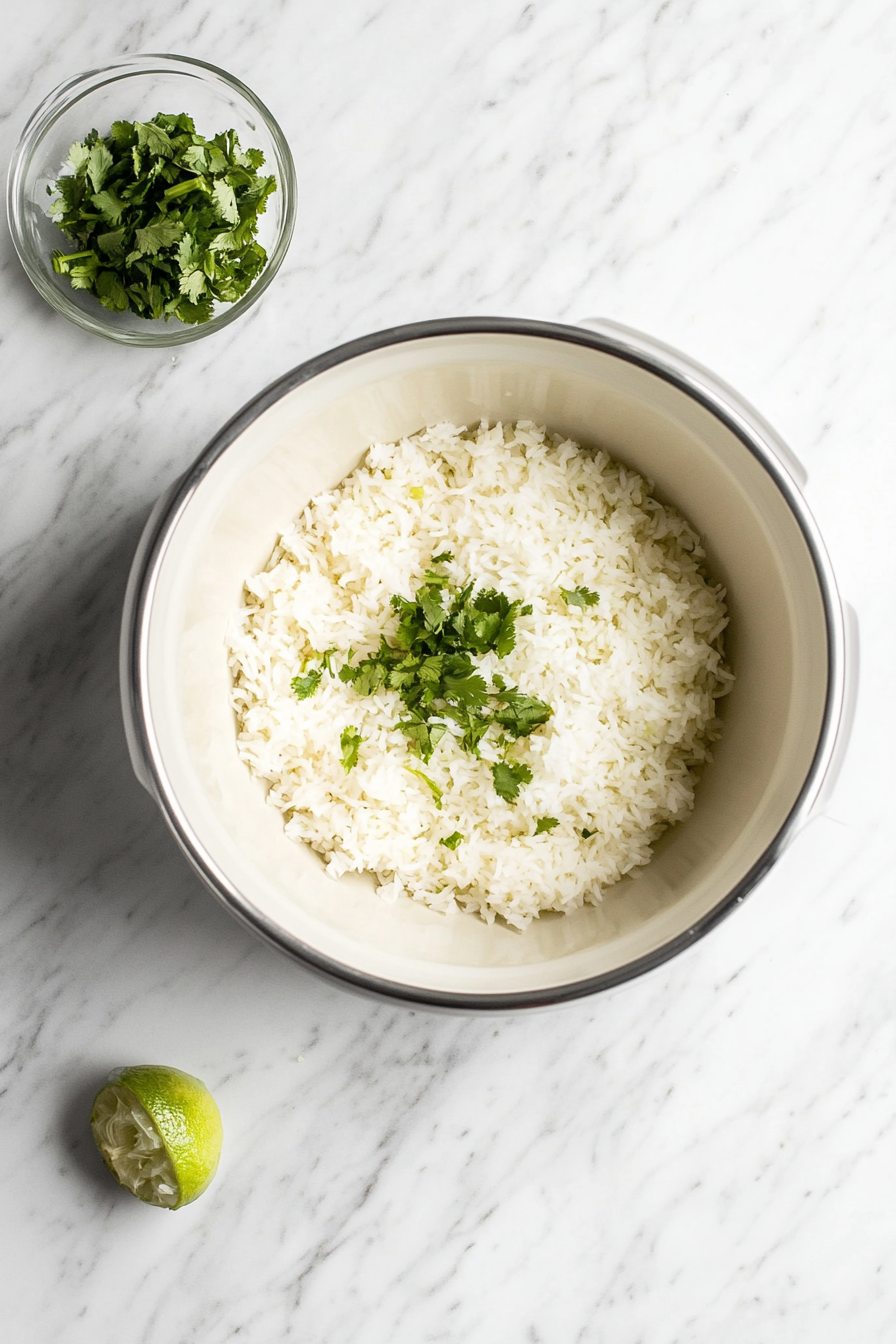 Top-down view of freshly cooked jasmine rice in a rice cooker, placed on a clean white marble countertop. The rice is fluffy and topped with chopped cilantro and a squeeze of lime juice. A small bowl with lime juice and cilantro sits nearby, enhancing the fresh flavors of the rice for a cilantro-lime rice base.