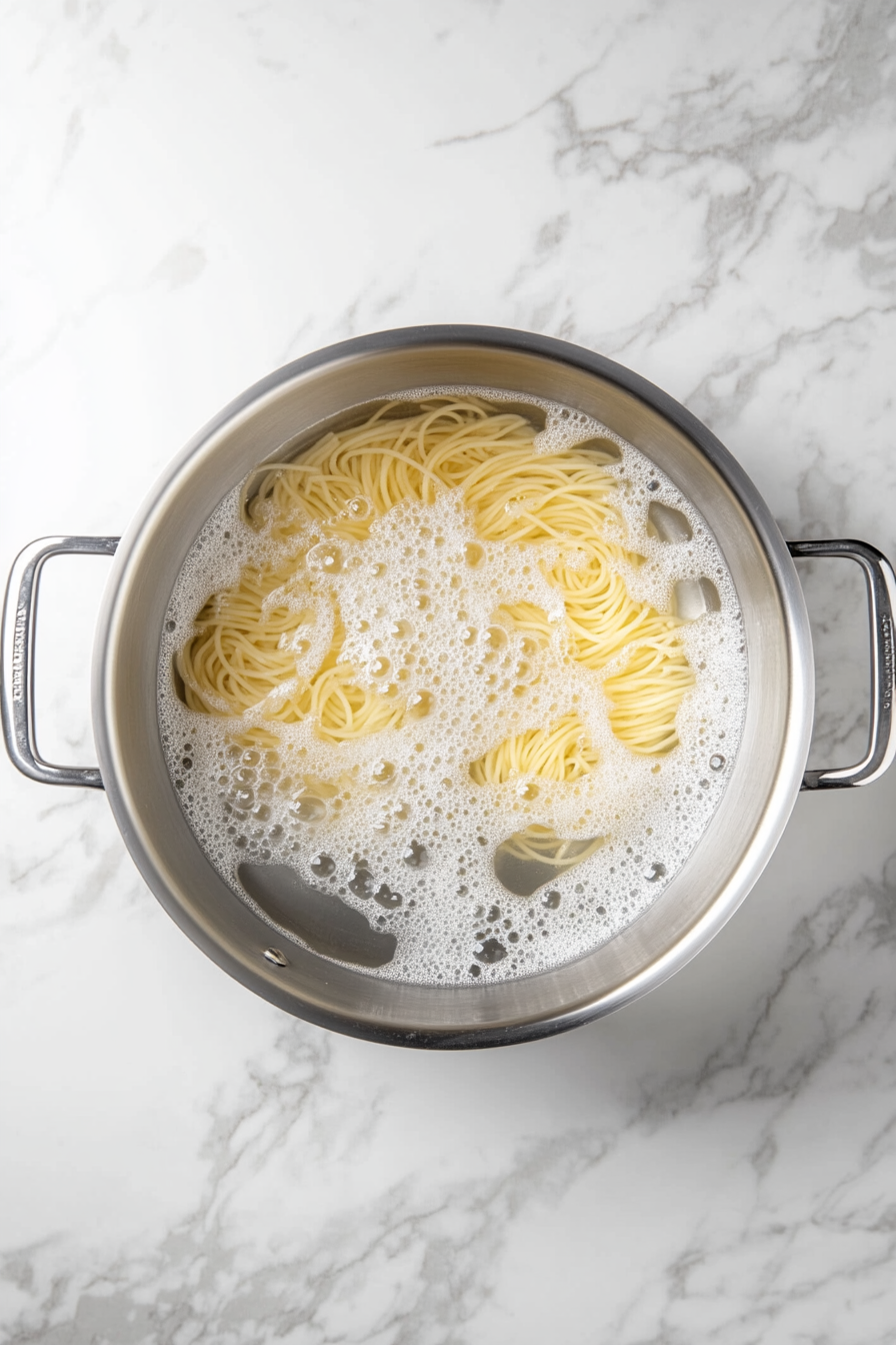 Top-down view showing spaghetti being cooked in a large stainless steel pot of salted boiling water. The pot is placed on a clean white marble cooktop, and the pasta is just beginning to soften. This step is essential for getting the perfect al dente texture before combining with the rich Alfredo sauce later in the recipe.
