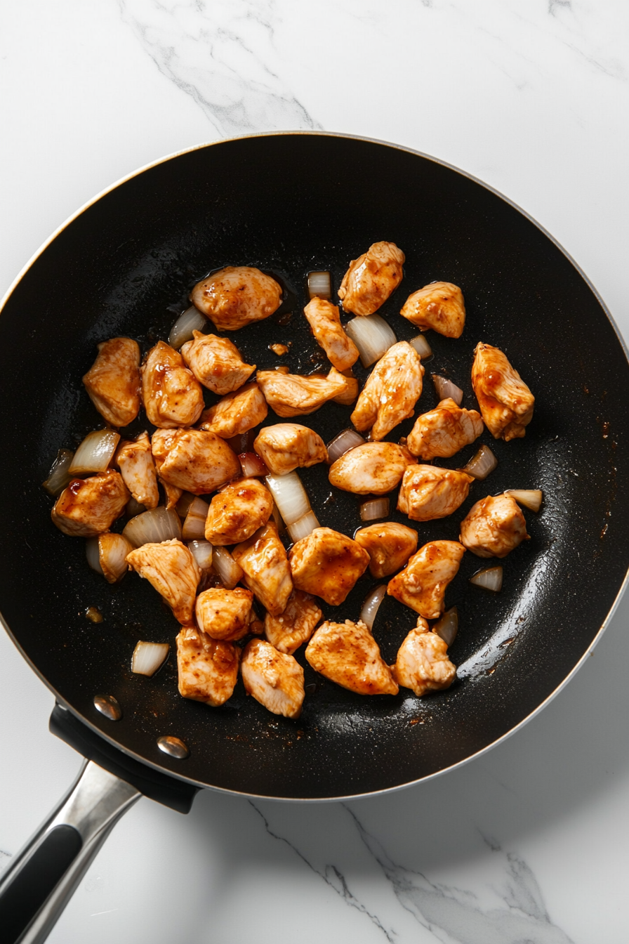 Top-down view of finely chopped onions being sautéed in a black non-stick skillet placed on a white marble cooktop. This step builds a fragrant base for the Alfredo sauce. The onions are just beginning to turn translucent, creating a flavor-rich foundation before adding the sauces and spices that elevate this creamy pasta recipe.