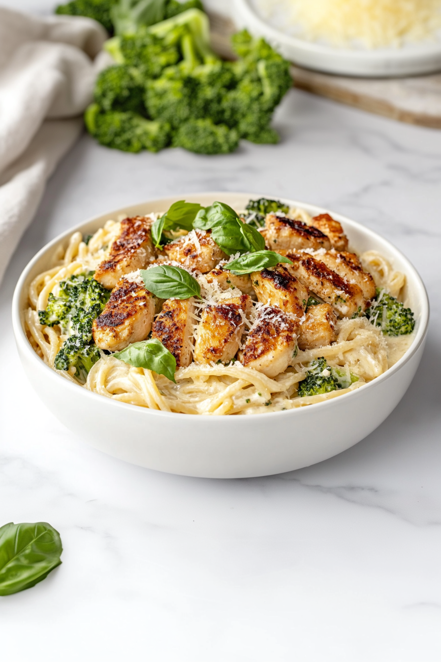 Top-down view of a comforting bowl of creamy Alfredo chicken pasta served in a white ceramic bowl on a white marble countertop. The dish features golden chicken chunks, creamy Alfredo-coated spaghetti, optional broccoli, and a generous sprinkle of shredded Parmesan cheese and fresh basil, creating a rich and visually inviting final presentation.