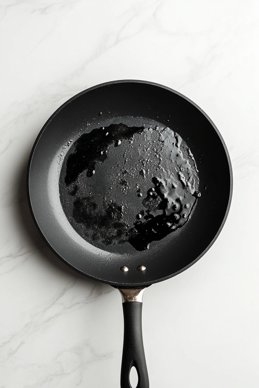 A mixture of dark soy sauce, honey, sriracha, and lemon juice is being simmered in a black non-stick skillet over a white marble countertop. The sauce is slightly thickening with small bubbles forming, indicating it is nearly ready. This bold and tangy stir fry sauce adds the signature flavor to the crispy chilli chicken.