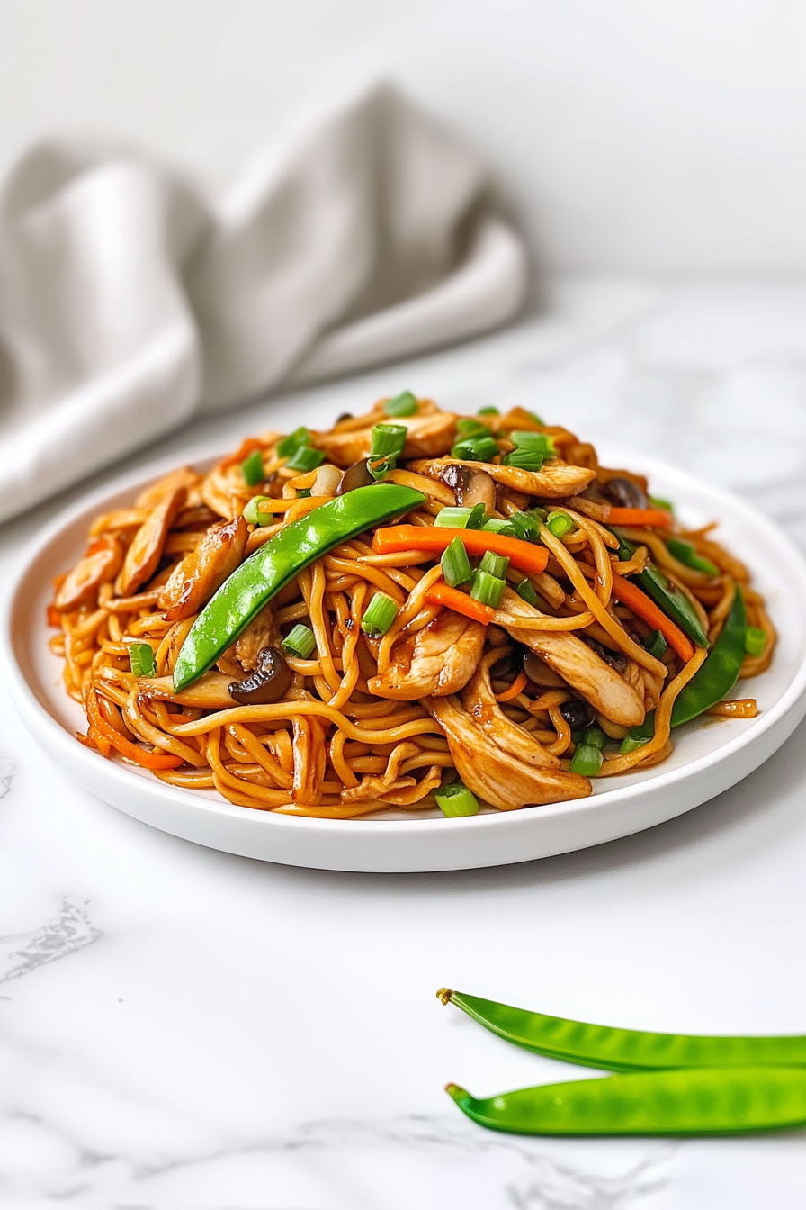 side view of a beautifully plated dish of stir-fried chicken with Hong Kong-style noodles, garnished with vibrant snap peas, carrots, mushrooms, mung bean sprouts, and scallions. The noodles are golden and slightly crispy, glistening with soy sauce and sesame oil. The clean and simple presentation over a white marble countertop adds elegance to the vibrant and fresh-looking meal, ready to be served with cucumber salad or grapefruit crush.