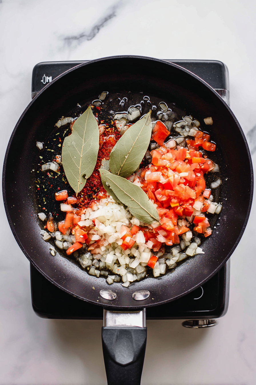 Heat oil in a skillet and sauté onion, tomato, garlic, chili, and bay leaves until fragrant.